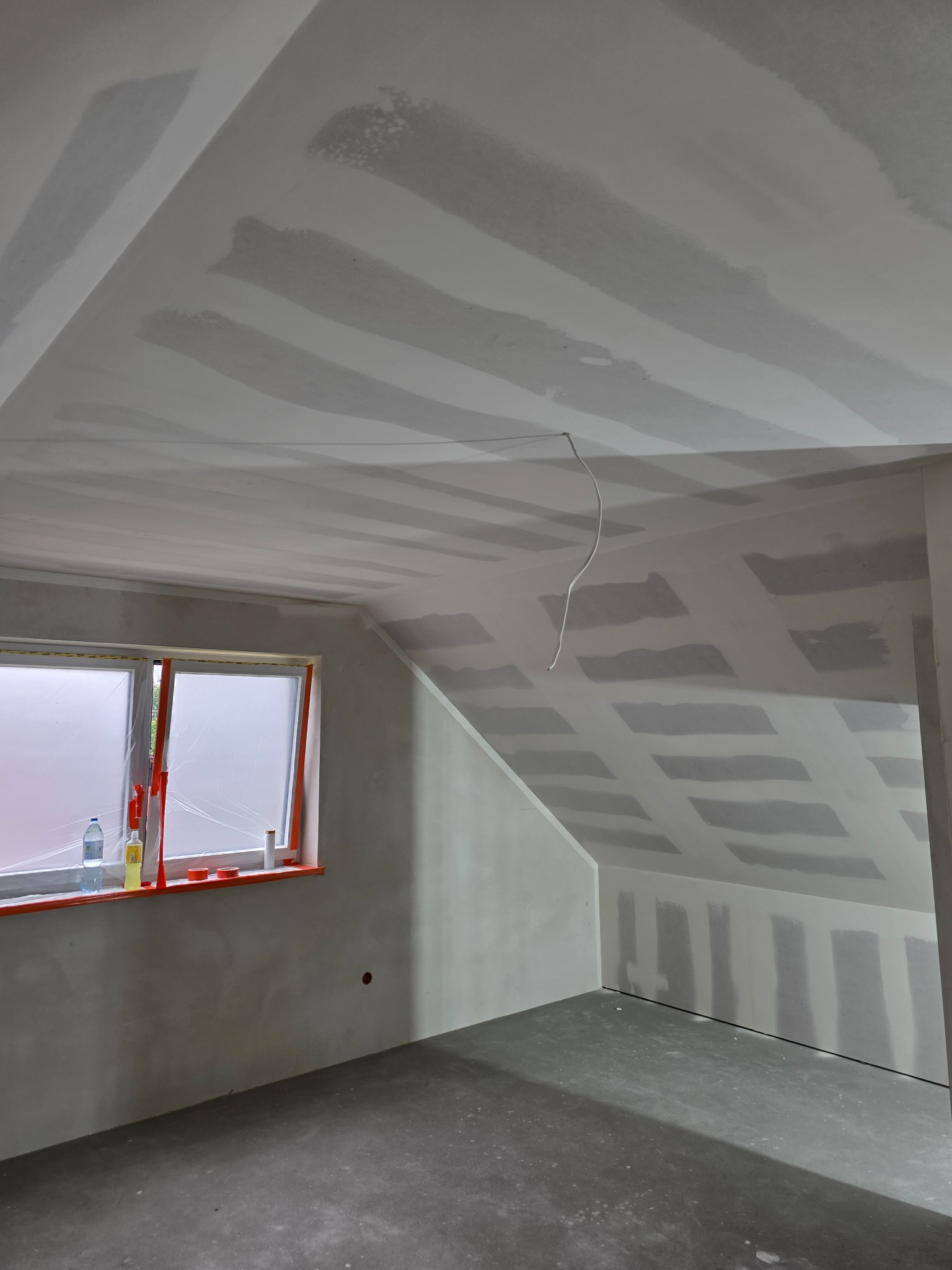 Poddasze po szpachlowaniu, widoczne łączenia płyt gipsowo-kartonowych na skosach i suficie. Okno zabezpieczone folią. W tle widoczny kabel elektryczny.