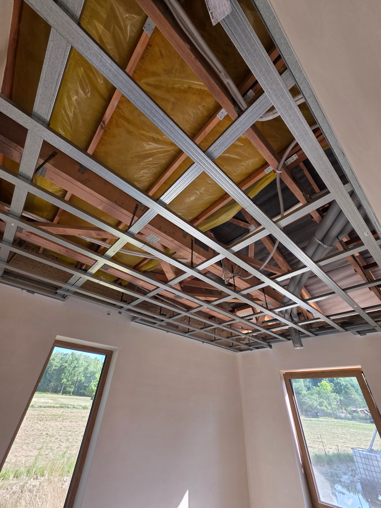 Sufit podwieszany w trakcie montażu: metalowa konstrukcja, izolacja z folii, drewniane elementy więźby dachowej. Widoczne okna dachowe i fragment ściany.
