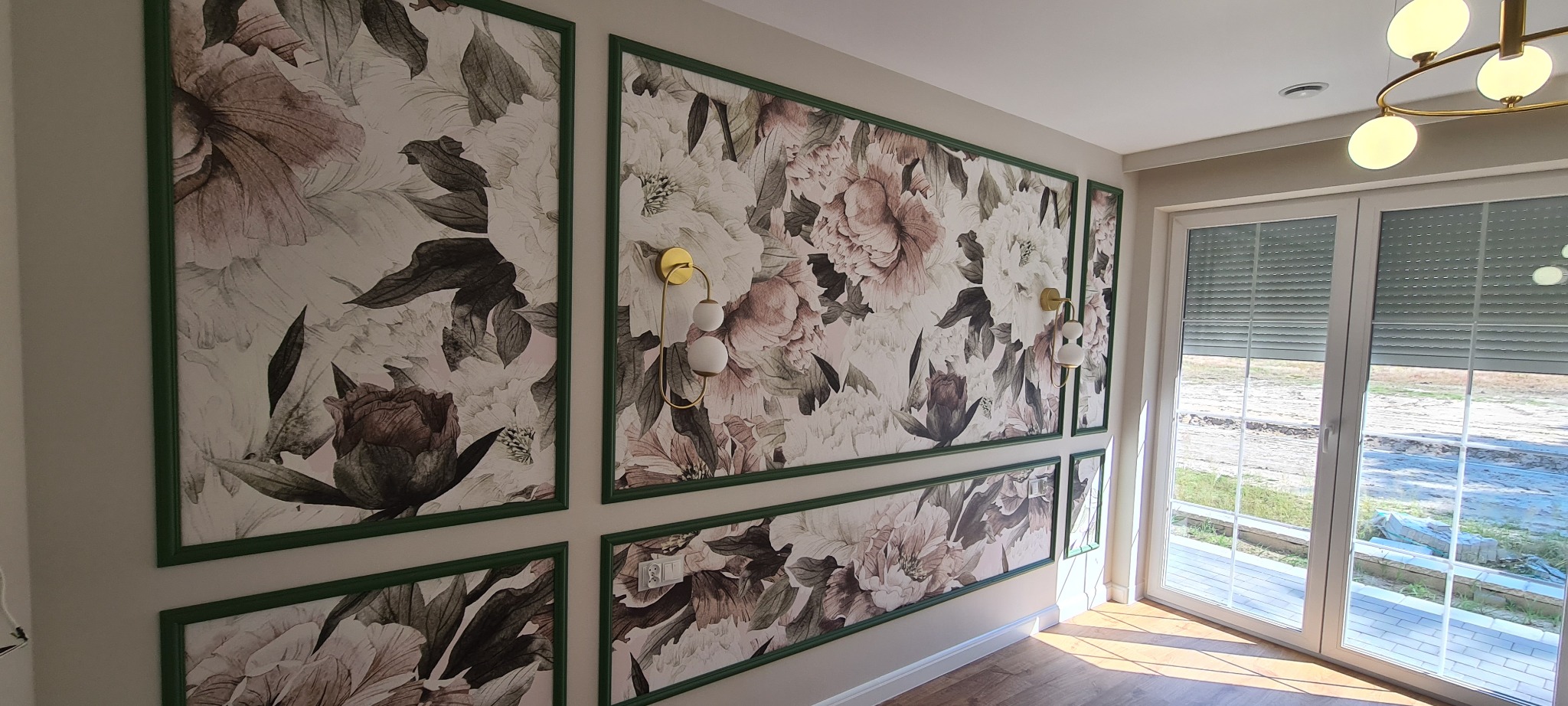 Tapeta w kwiaty z zielonymi ramkami na ścianie, oświetlenie ścienne w kolorze złota, widok na okno z roletami. Dekoracyjne wykończenie wnętrza.