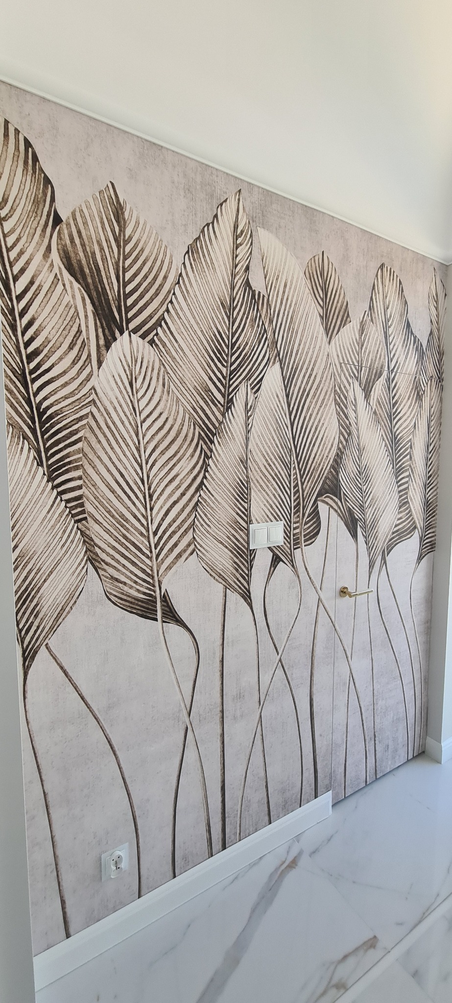 Beżowa tapeta w liście na ścianie z włącznikiem i gniazdkiem, biała podłoga z marmuru. Wykończenie wnętrza w neutralnych barwach.