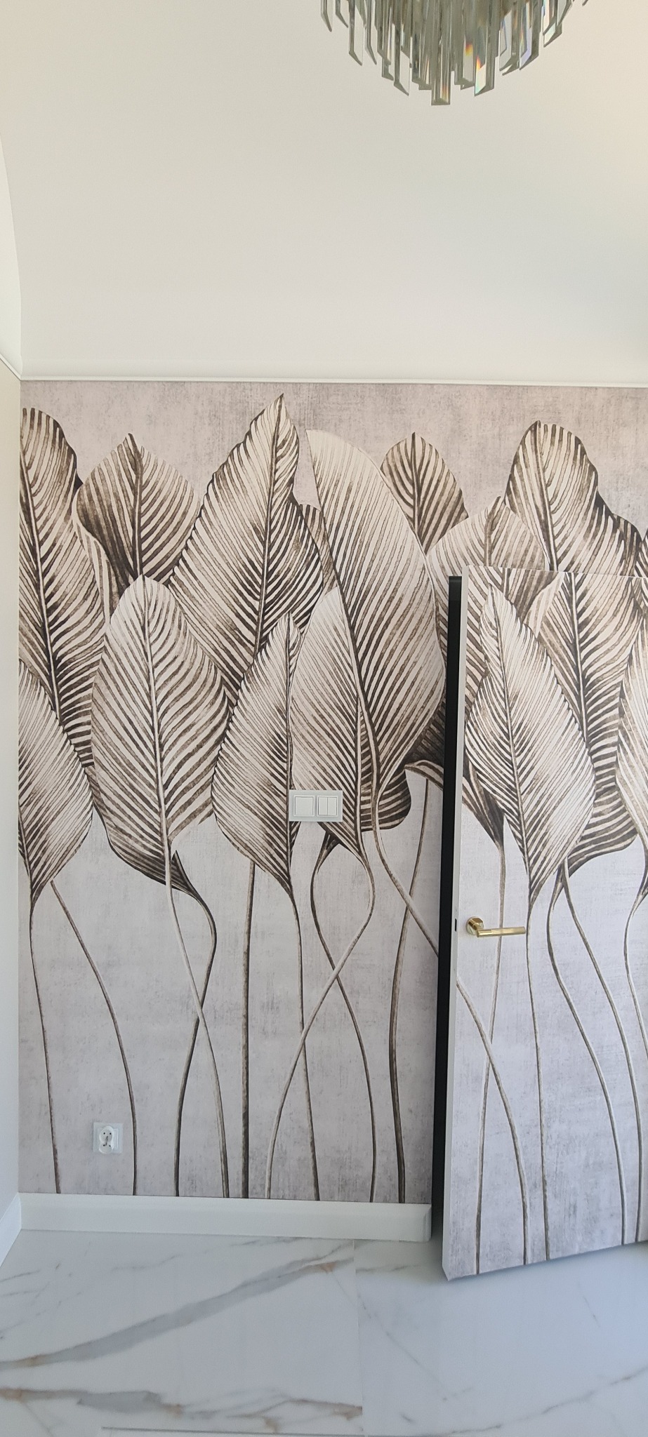 Tapeta w liście na ścianie w jasnym wnętrzu, widoczny fragment drzwi z złotą klamką, marmurowa podłoga i nowoczesny żyrandol. Neutralna kolorystyka.