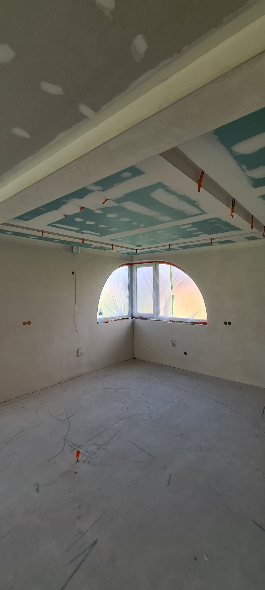 Pokój w trakcie wykończenia: szpachlowana ściana, sufit z płyt gipsowo-kartonowych z dekoracyjnym wzorem, okno w kształcie łuku zabezpieczone folią malarską.