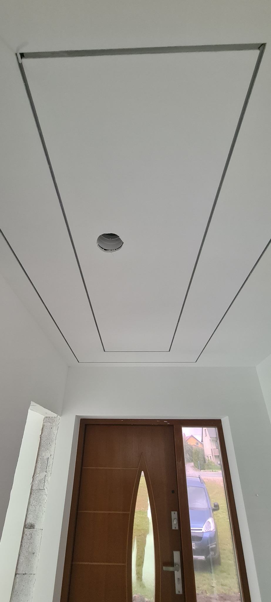 Biały sufit z dekoracyjnymi, szarymi listwami tworzącymi geometryczny wzór. Widoczny otwór na oświetlenie i brązowe drzwi wejściowe z szybą.