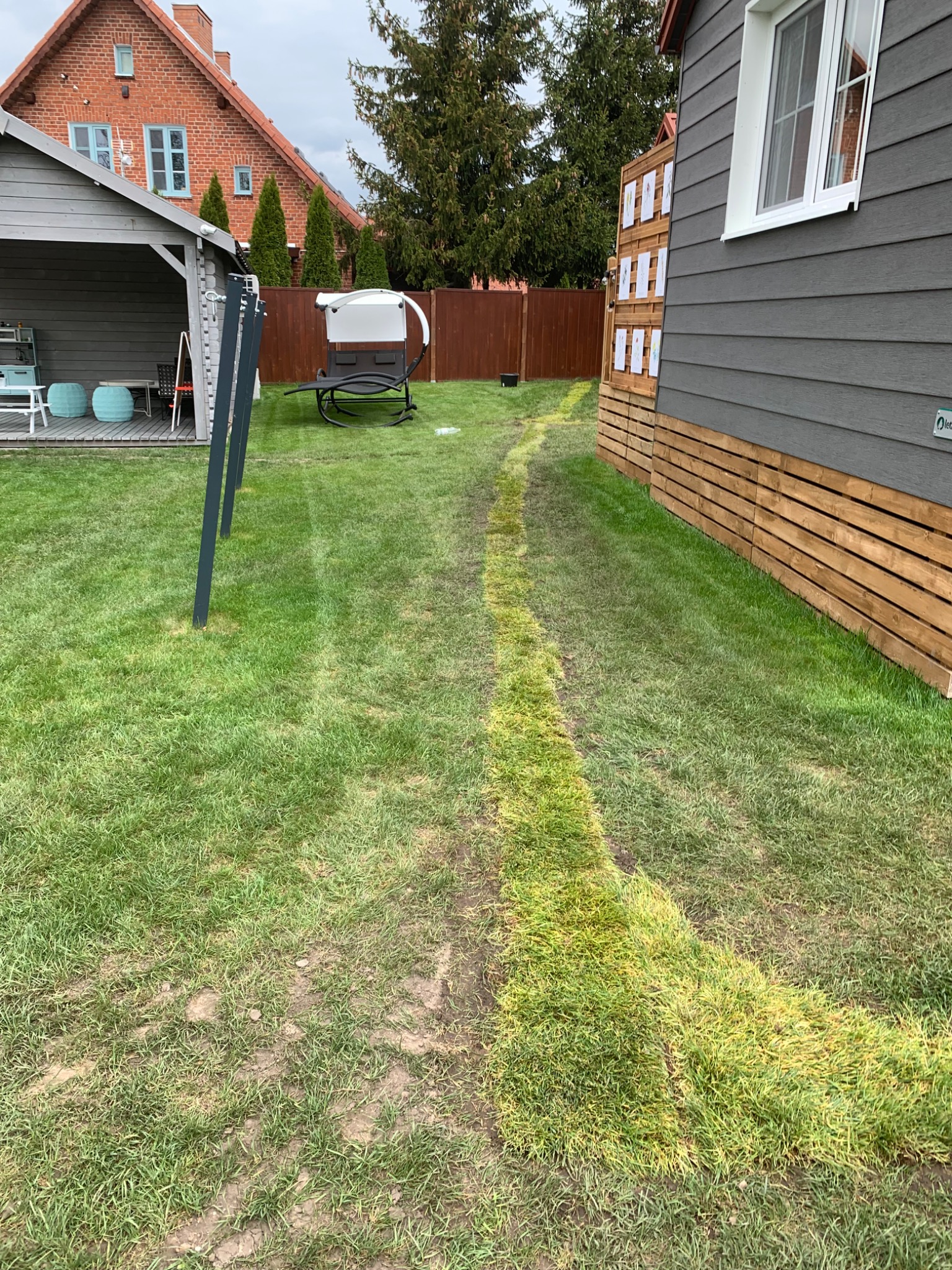 Świeżo założony trawnik z widoczną ścieżką po przejeździe maszyny, obok drewnianego domku z szarą elewacją i elementami z drewna, w tle domek narzędziowy i dom z czerwonej cegły.