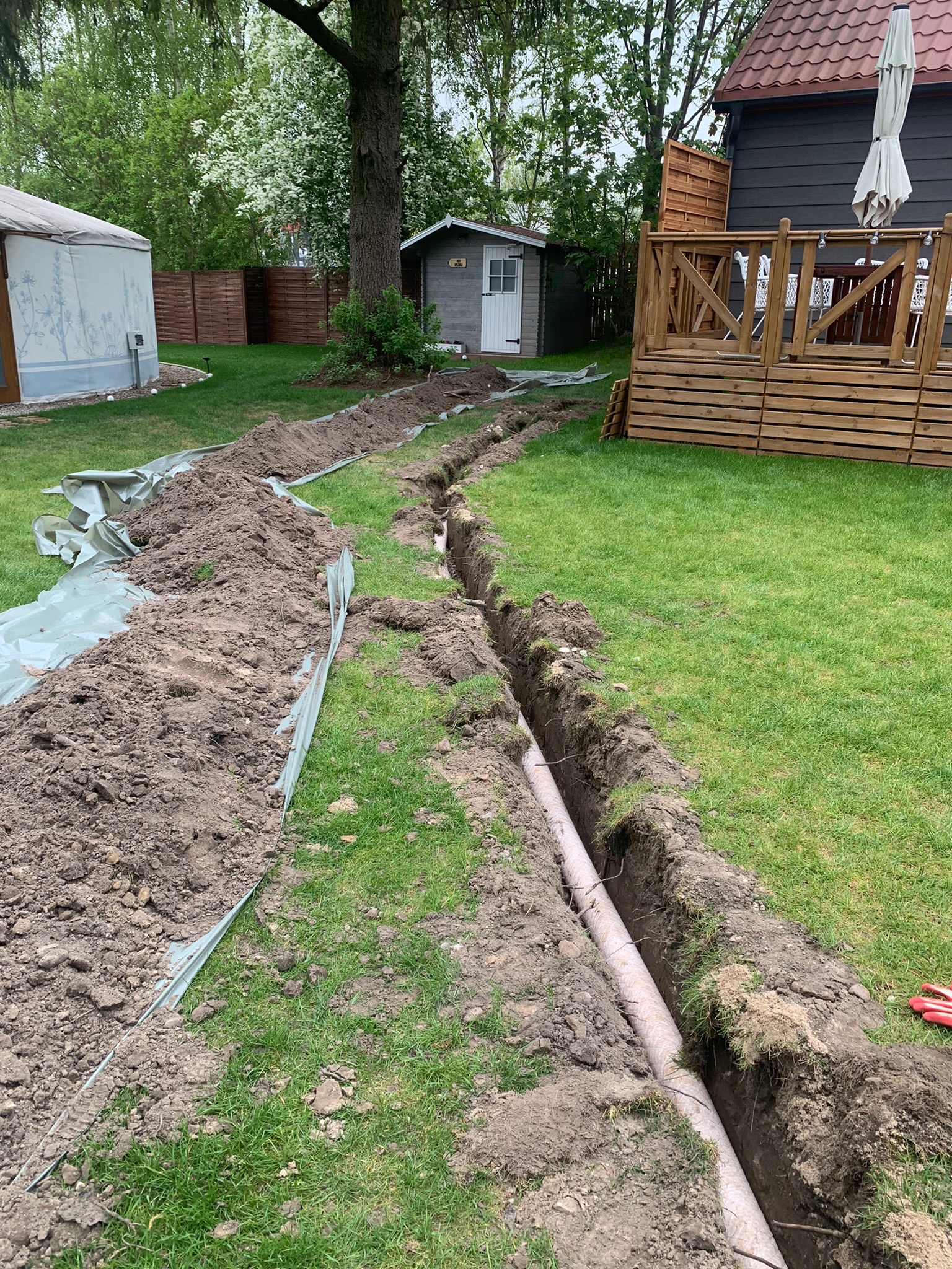 Wykopany rów pod instalację nawadniającą w ogrodzie, widoczne ułożone rury, ziemia zabezpieczona folią, w tle domek narzędziowy i taras.
