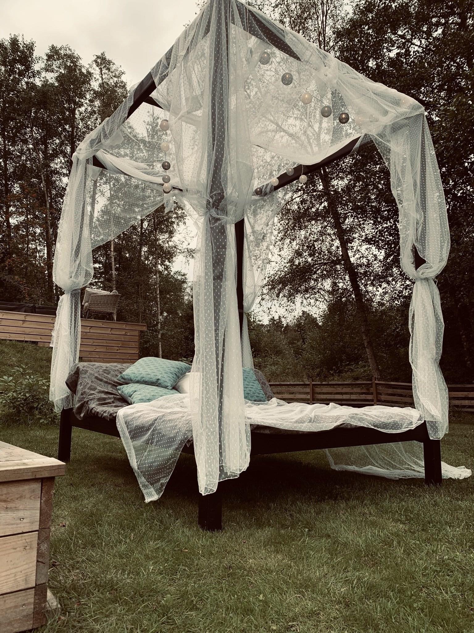 Zadaszone łóżko ogrodowe z baldachimem z białej tkaniny w kropki, oświetlone girlandą z kul, ustawione na trawniku w ogrodzie.