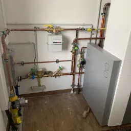 Instalacja grzewcza z kotłem Wolf, rurami miedzianymi, zaworami i licznikiem gazu na białej ścianie w pomieszczeniu z brązowymi płytkami podłogowymi.