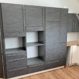 Nowoczesna zabudowa kuchenna z szarymi frontami imitującymi kamień, z długimi, srebrnymi uchwytami, połączona z białą zabudową i drewnianym blatem.