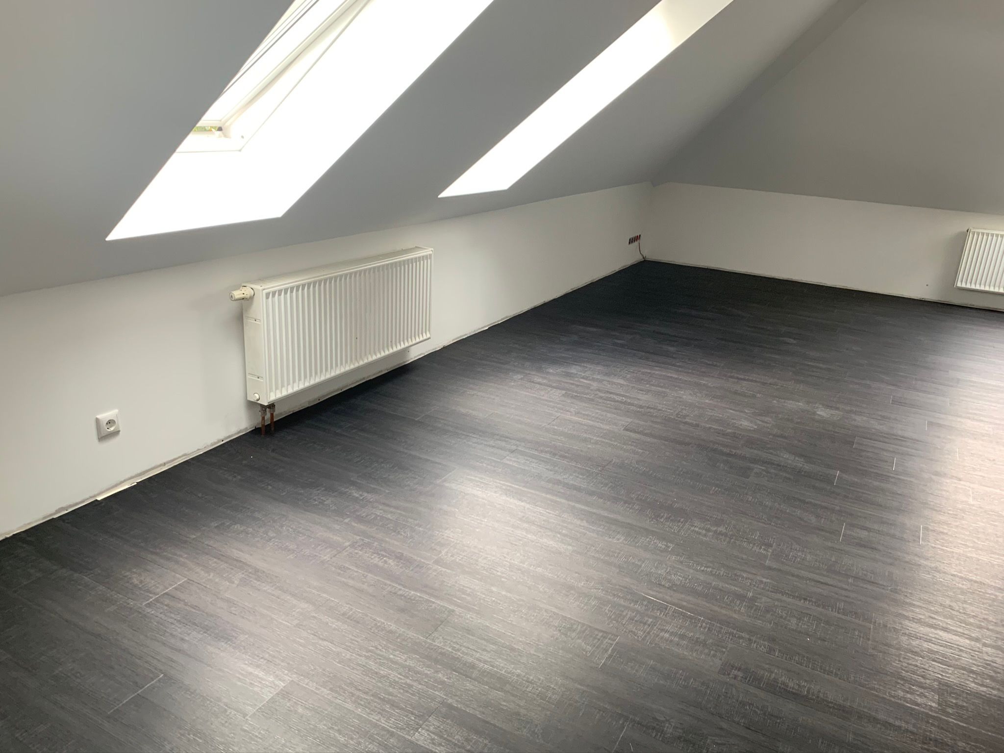 Poddasze z trzema oknami dachowymi, grzejnikiem i ciemną podłogą imitującą drewno, widoczne gniazdko elektryczne i niedokończone listwy przypodłogowe.