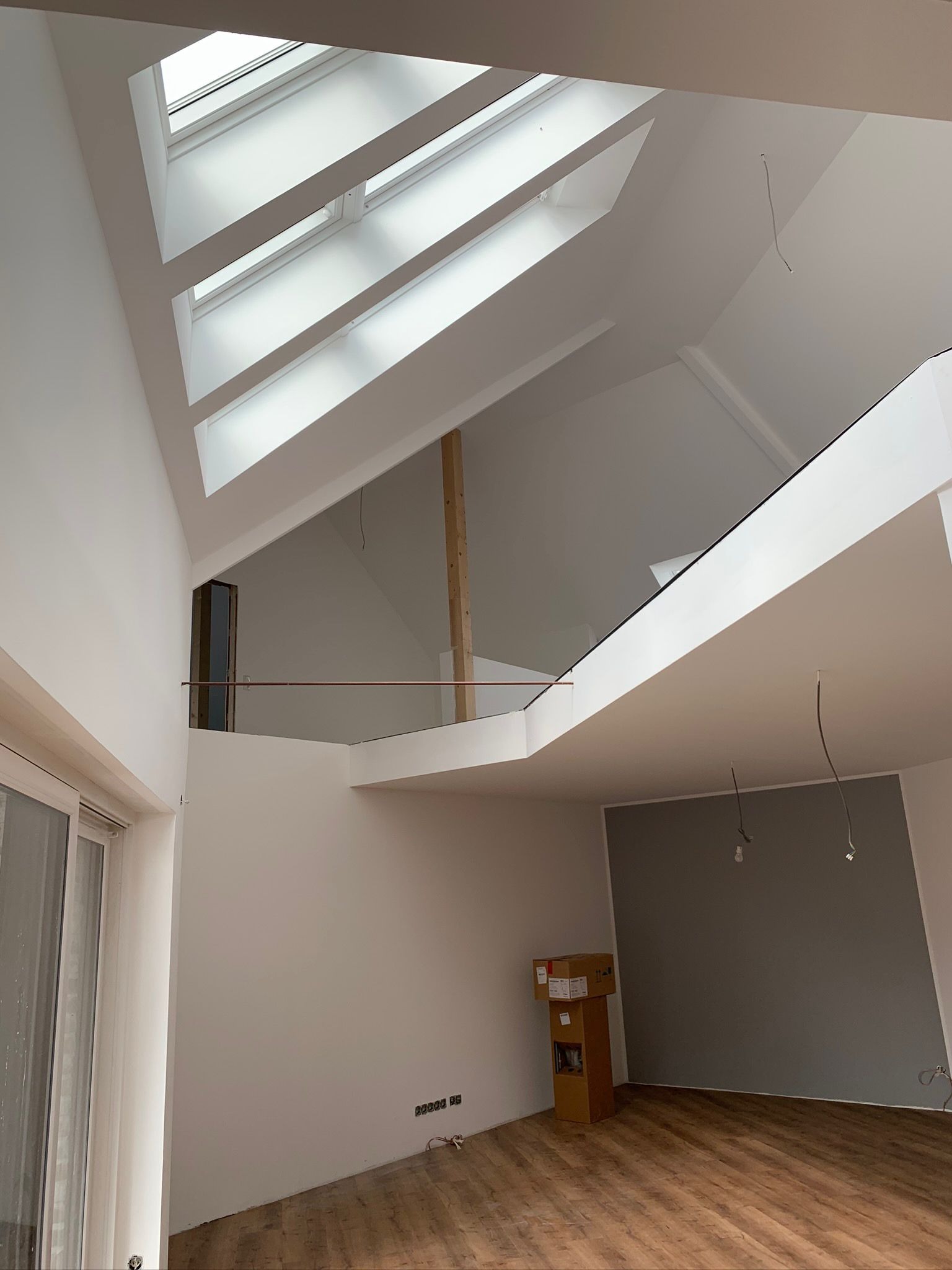 Wysokie wnętrze z antresolą i oknami dachowymi, świeżo pomalowane na biało, z drewnianą podłogą i wystającymi kablami od oświetlenia, kartony w rogu pokoju.