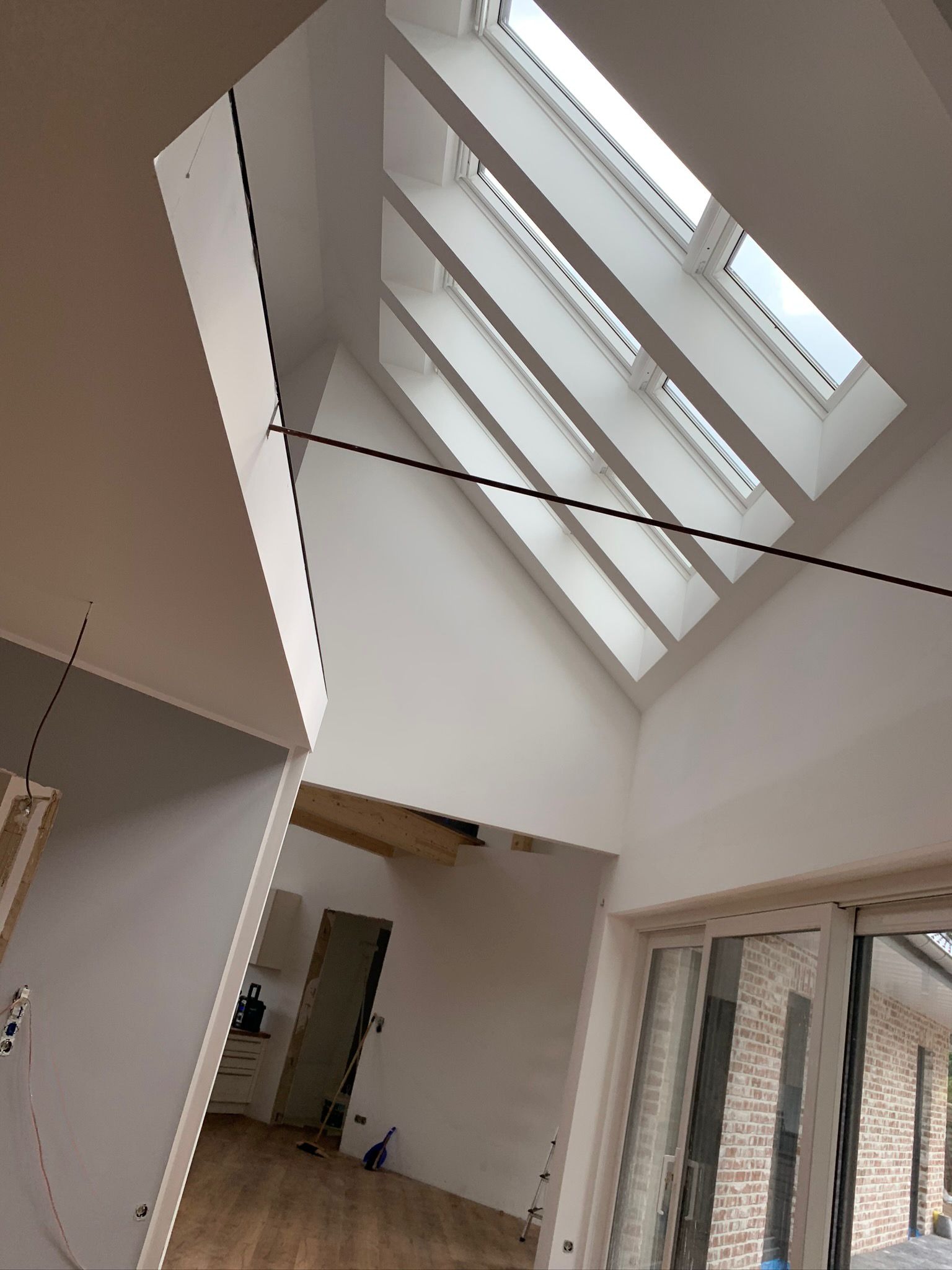 Wysokie wnętrze z oknami dachowymi w skośnym dachu, ściany pomalowane na biało, widoczne elementy instalacji elektrycznej i fragmenty podłogi w trakcie wykańczania.