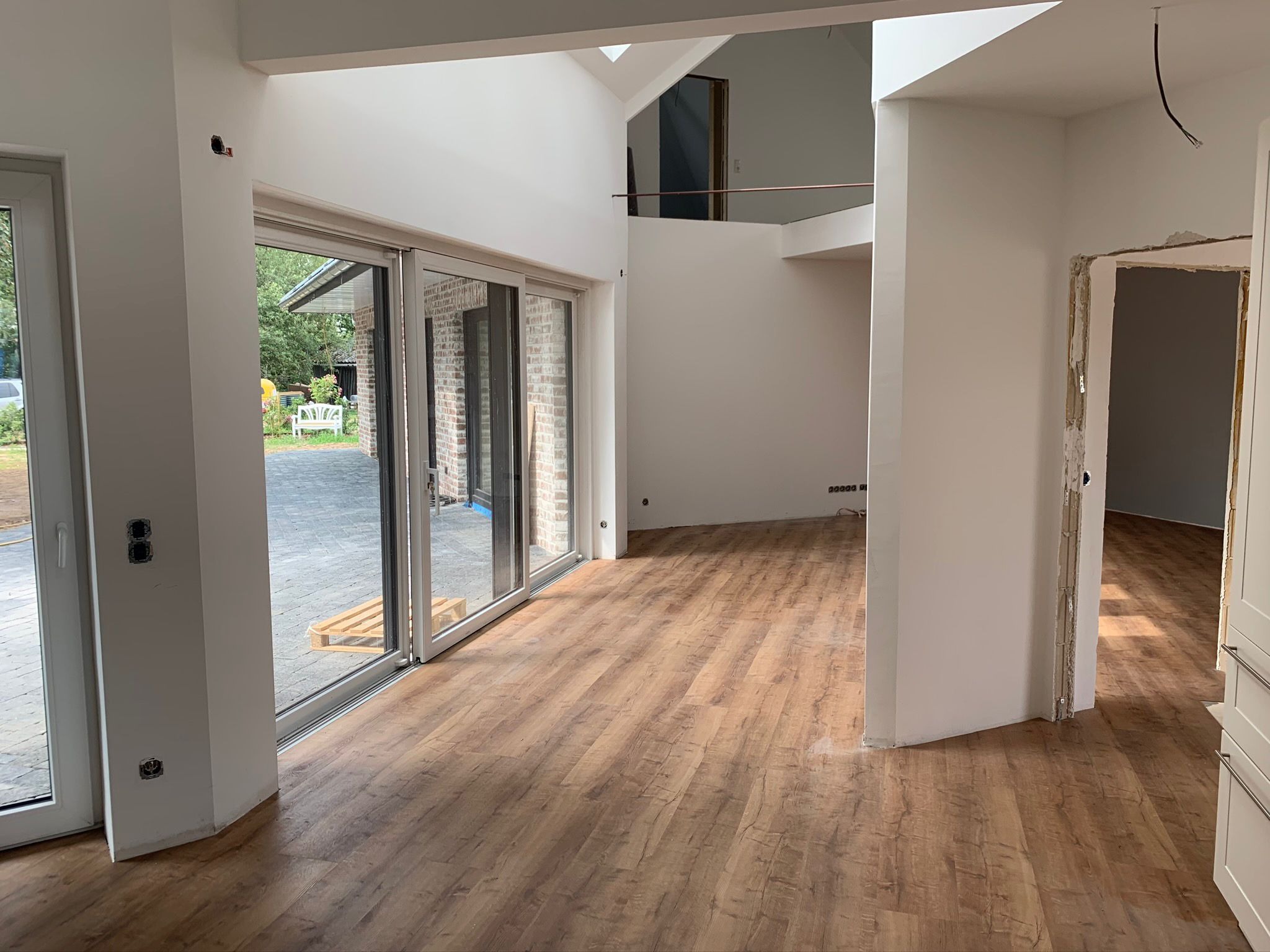 Wnętrze domu w trakcie wykańczania, widoczne nowe panele podłogowe, białe ściany z otworami na gniazdka elektryczne, duże przesuwne okna z widokiem na ogród i dwupoziomowa przestrzeń z elementami...