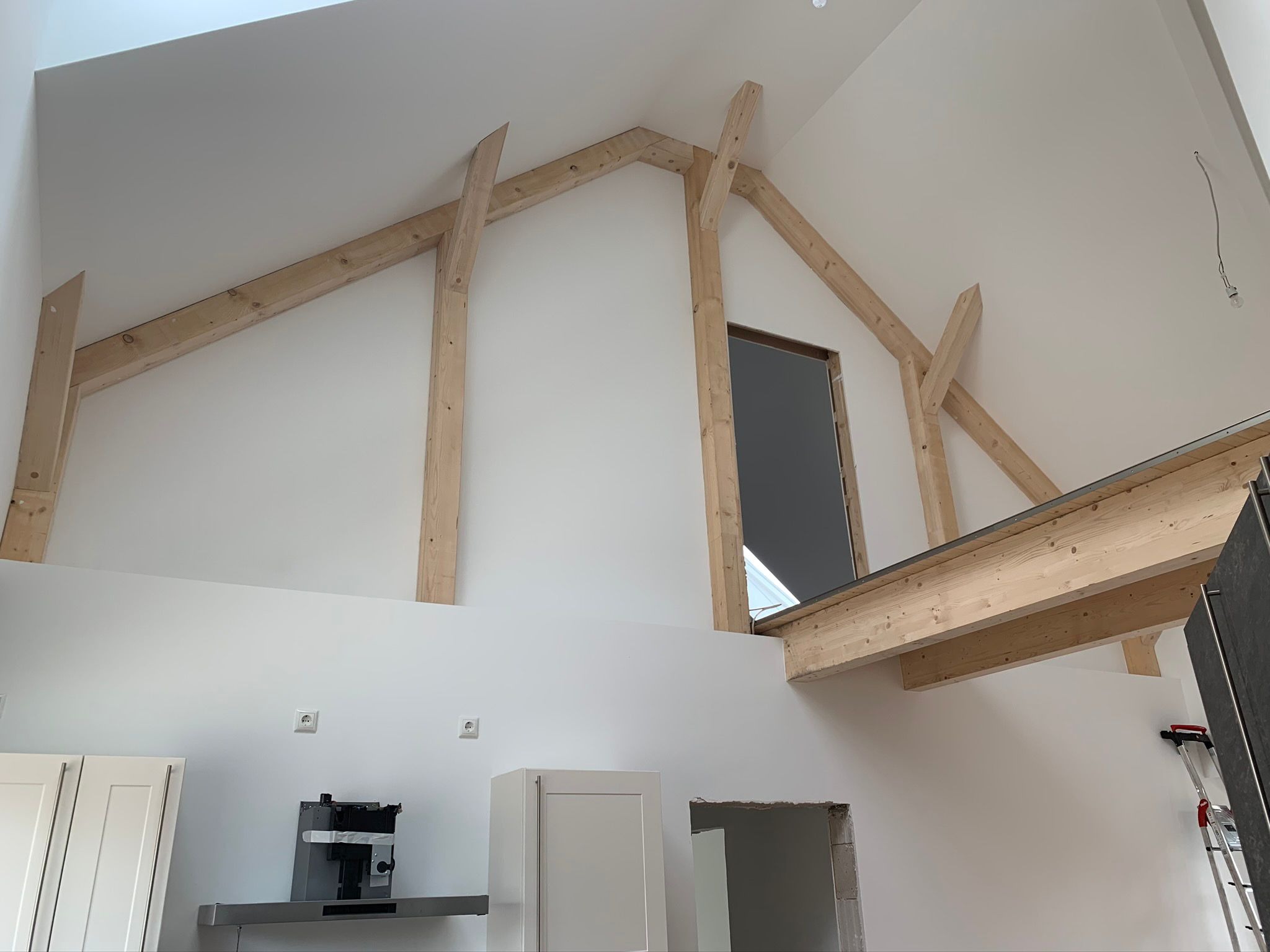 Wnętrze domu w trakcie budowy, widoczna drewniana konstrukcja więźby dachowej, białe ściany, elementy zabudowy kuchennej i drabina