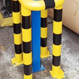 Ochronna konstrukcja z rur stalowych w kolorze ż&oacute;łto-czarnym, zabezpieczająca niebieski słup, zamocowana na betonowej posadzce przy pomocy śrub.