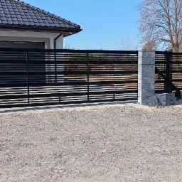 Nowoczesna, przesuwna brama wjazdowa z czarnymi, poziomymi profilami i szarymi, betonowymi słupkami, na tle domu z ciemną dachówką i niebieskiego nieba.