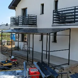 Nowoczesny dom z czarnymi balustradami balkonowymi i zadaszeniem nad wejściem, widoczne narzędzia i materiały budowlane wskazują na trwające prace wykończeniowe.