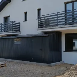 Nowoczesny, antracytowy schowek narzędziowy z metalu, przylegający do ściany budynku z balkonami z czarnymi balustradami, widoczne ślady prac ziemnych i materiały budowlane.