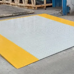 Metalowa rampa najazdowa z blachy ryflowanej, pomalowana na szaro z żółtymi krawędziami, przymocowana do betonowej posadzki, w tle palety i czerwona gaśnica.