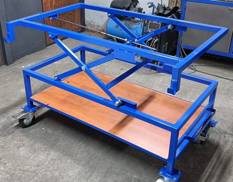 Niebieska, stalowa rama podnośnika warsztatowego na kółkach z drewnianą platformą, widoczny kompresor w tle.