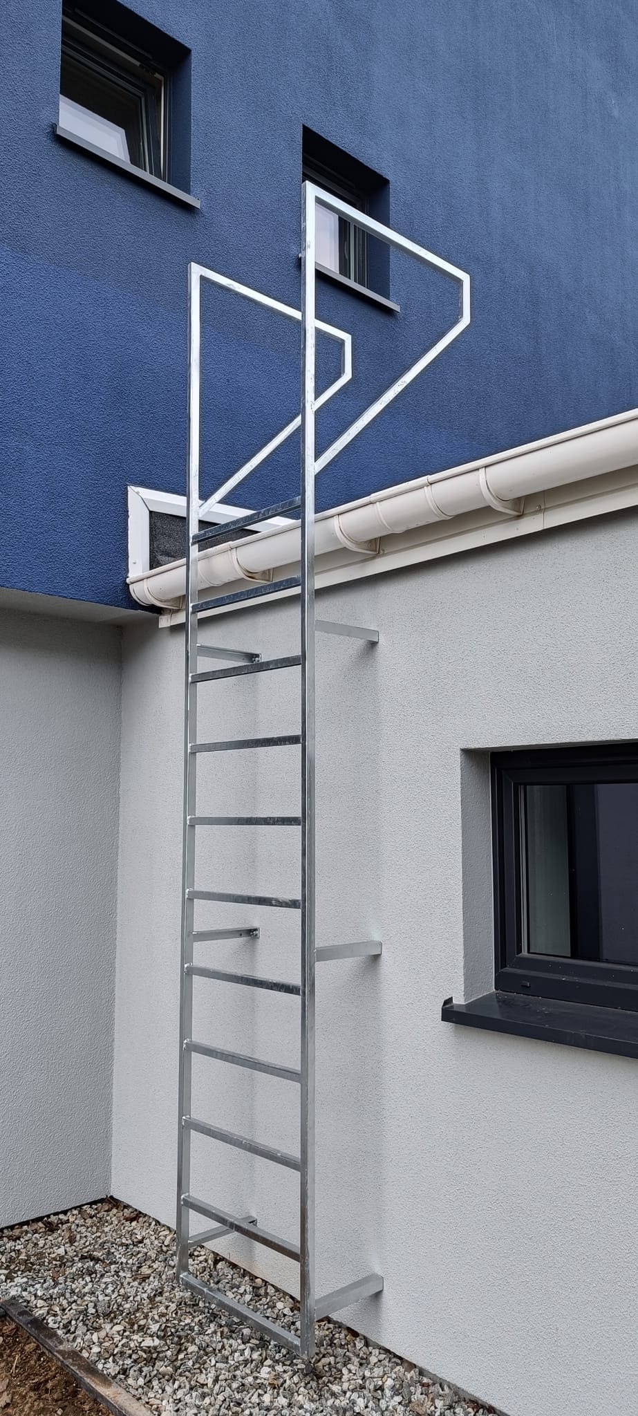 Zewnętrzna, metalowa drabina ewakuacyjna przymocowana do ściany budynku w Koszalinie, widok z dołu, na dole wysypany żwir.