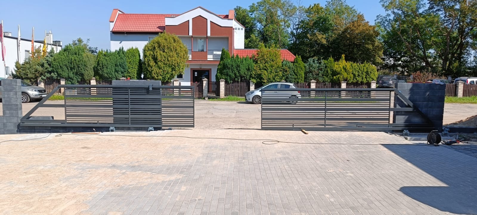 Nowoczesna, przesuwna brama wjazdowa z poziomymi panelami, zamontowana przed posesją z budynkiem mieszkalnym i zadbanym ogrodem, widoczne ślady montażu.
