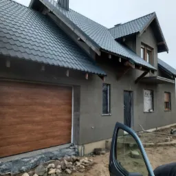 Surowy dom jednorodzinny z szarą elewacją w trakcie prac wykończeniowych, drewniana brama garażowa, dach pokryty blachodachówką w kolorze grafitowym, okna zabezpieczone folią, widoczne...