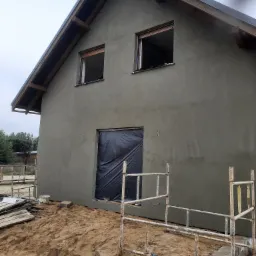 Surowy, nieotynkowany budynek jednorodzinny z drewnianą konstrukcją dachu, zabezpieczonym otworem drzwiowym folią i rozstawionym rusztowaniem.