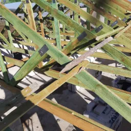 Wnętrze budowy domu w trakcie montażu więźby dachowej, widoczne drewniane elementy konstrukcyjne w odcieniach zieleni i brązu, połączone metalowymi płytkami, z widokiem na niebo w tle.