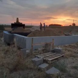 Wylany fundament z izolacją, zasypany piaskiem, w tle koparka i sylwetki dwóch mężczyzn pracujących przy zachodzącym słońcu.