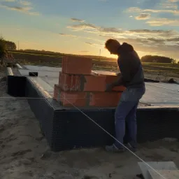 Pracownik w szarym ubraniu układa cegły na fundamencie domu, widok na zachodzące słońce i pole w tle.