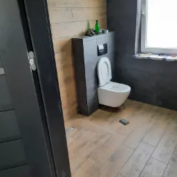 Nowoczesna toaleta podwieszana z deską sedesową w kolorze białym, zamontowana w łazience wykończonej płytkami imitującymi drewno i szarym gresem. Na zabudowie stelaża widoczna zielona butelka...