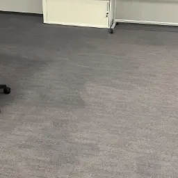 Szara, wzorzysta podłoga w biurze, z widocznym fragmentem nogi krzesła obrotowego, szafką z dokumentami i tablicą informacyjną na kółkach.