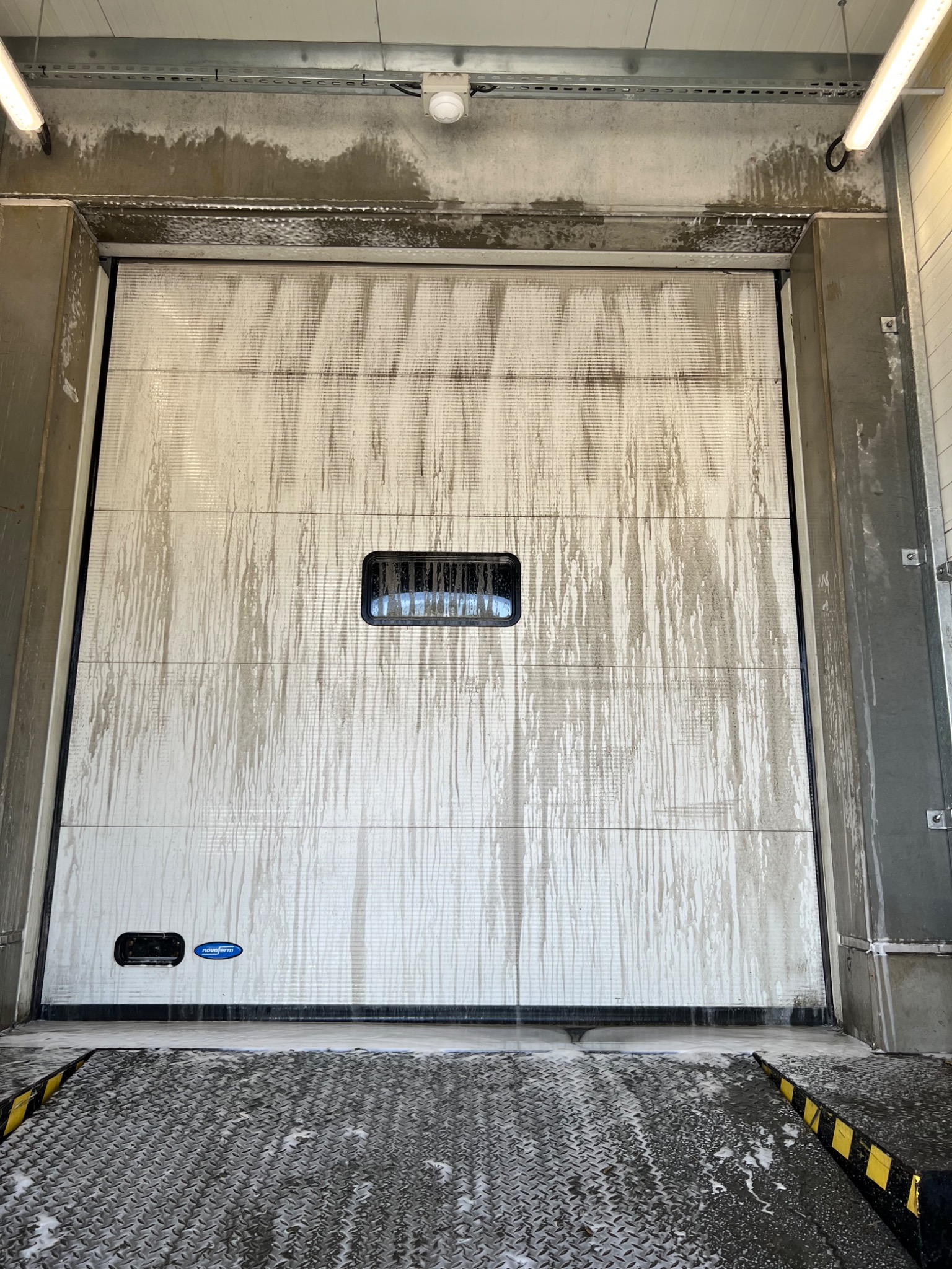 Brama przemysłowa z widocznymi śladami mycia pod ciśnieniem, z zaciekami i pianą na posadzce z blachy ryflowanej.