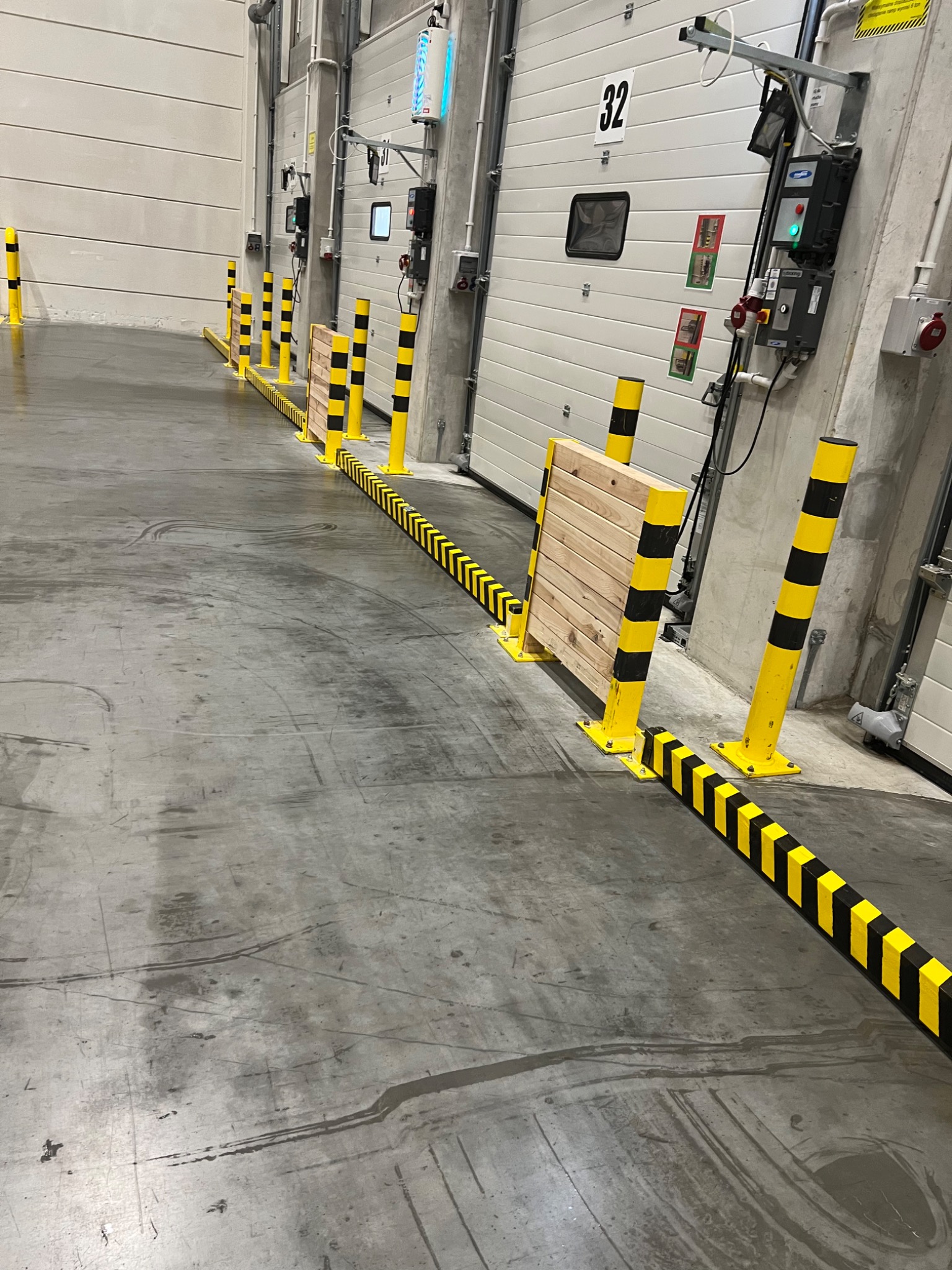 Wnętrze hali magazynowej z betonową posadzką, zabezpieczoną żółto-czarnymi słupkami i barierami ochronnymi przy bramach wjazdowych.