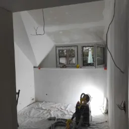 Pokój na poddaszu w trakcie prac wykończeniowych, widoczna szlifierka do gładzi gipsowych oraz zakryta folią podłoga.