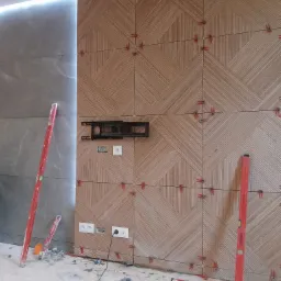 Ściana wyłożona płytkami z wzorem jodełki, z widocznymi krzyżykami dystansowymi i poziomnicami opartymi o ścianę, gniazdka elektryczne i uchwyt do telewizora zamontowane.