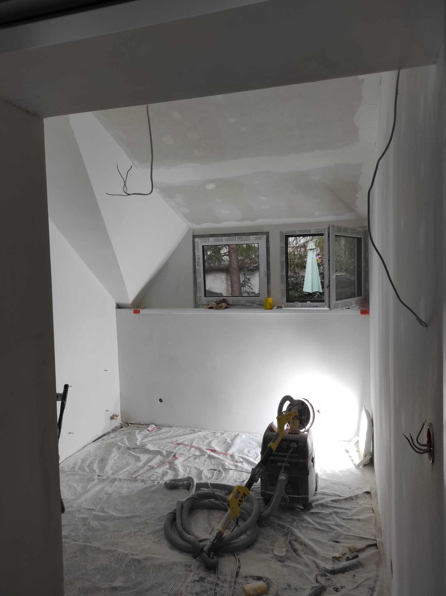 Pokój na poddaszu w trakcie prac wykończeniowych, widoczna szlifierka do gładzi gipsowych oraz zakryta folią podłoga.