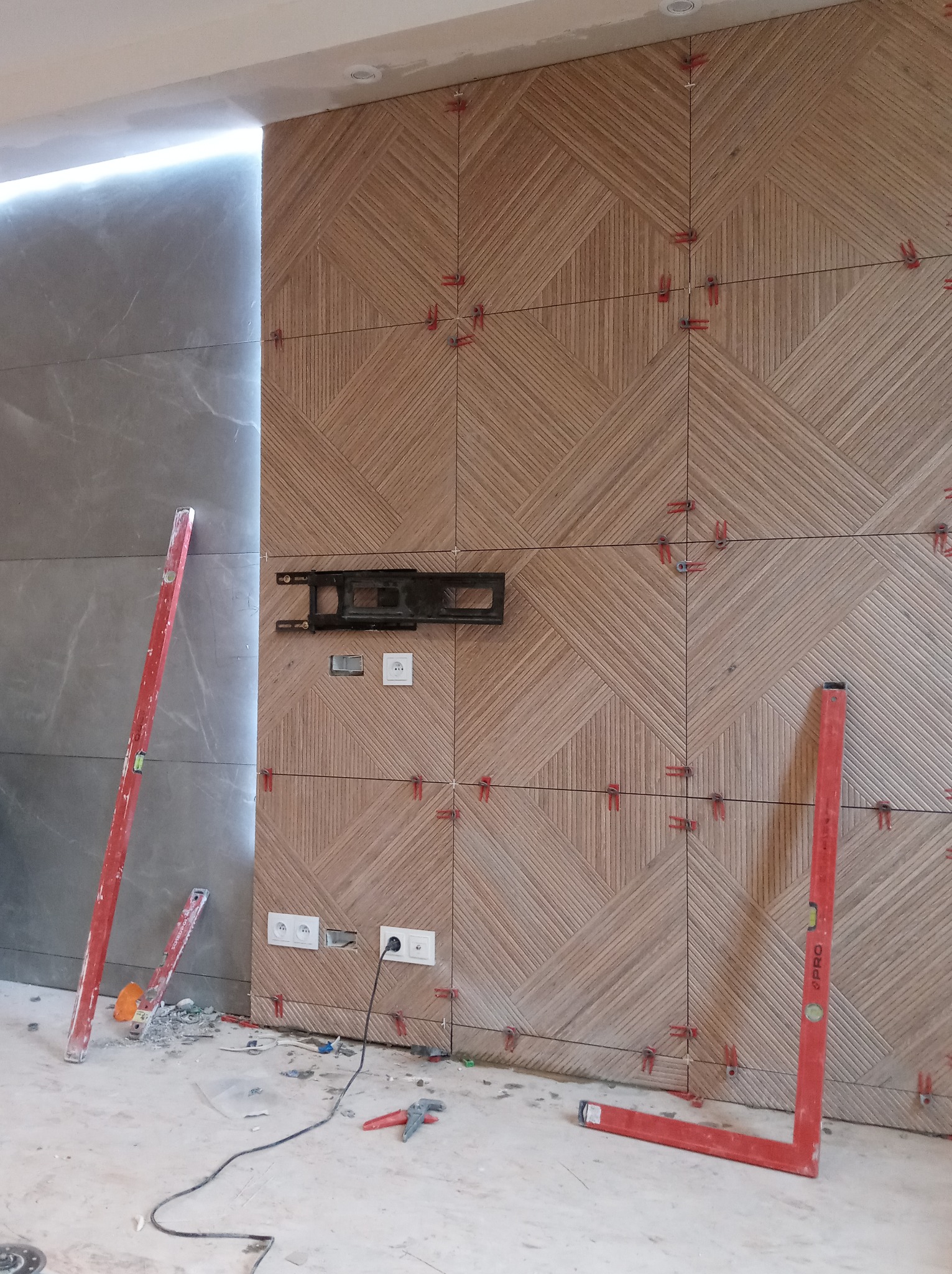 Ściana wyłożona płytkami z wzorem jodełki, z widocznymi krzyżykami dystansowymi i poziomnicami opartymi o ścianę, gniazdka elektryczne i uchwyt do telewizora zamontowane.