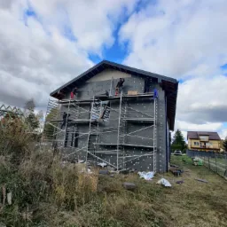 Dom jednorodzinny w trakcie prac związanych z izolacją termiczną elewacji, widoczne rusztowania i ekipa montująca płyty izolacyjne na ścianach budynku, w tle błękitne niebo z chmurami.
