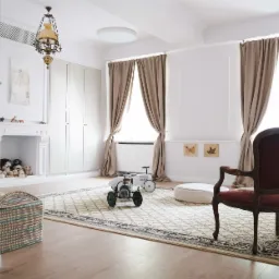 Jasne wnętrze pokoju dziecięcego z drewnianą podłogą, dywanem w kremowe wzory, zabawkami, fotelikiem w stylu retro, zasłonami w kolorze beżowym i dekoracyjnym kominkiem z pluszakami.