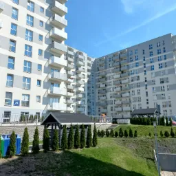 Nowoczesny kompleks apartamentowy z jasną elewacją, balkonami i zagospodarowanym terenem zielonym z altaną, drzewkami i kolorowymi pojemnikami na odpady.