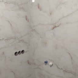 VPBud - Ściana w łazience wykończona płytkami imitującymi marmur, widoczne otwory na baterię, przyłącza wodne i odpływ w podłodze, etap prac wykończeniowych.