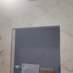 Ściana wyłożona jasnymi płytkami imitującymi marmur z widocznym otworem drzwiowym w trakcie prac wykończeniowych, z fragmentem drewnianej konstrukcji i rękawicami roboczymi w tle.