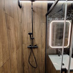 VPBud - Nowoczesna kabina prysznicowa z deszczownicą i panelem ściennym imitującym drewno, oświetlone lustro z umywalką w tle.