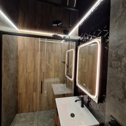 VPBud - Nowoczesna łazienka z kabiną prysznicową typu walk-in, umywalką nablatową na szafce z drewnopodobnym dekorem, podświetlanymi lustrami i czarną armaturą.