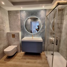VPBud - Nowoczesna łazienka we Wrocławiu z szarymi płytkami imitującymi marmur, podwieszana toaleta, umywalka z szafką w kolorze granatowym i okrągłe lustro z podświetleniem LED oraz kabina prysznicowa ze...
