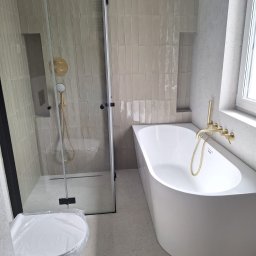 VPBud - Nowoczesna łazienka z beżowymi, pionowo ułożonymi płytkami, kabiną prysznicową typu walk-in z czarną ramą, złotą armaturą i białą wanną.