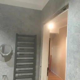 Szara łazienka z grzejnikiem drabinkowym i okrągłym lustrem, widok na korytarz z drewnianą podłogą przez otwarte drzwi bez ościeżnicy.