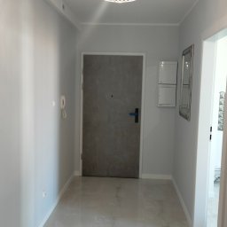 VPBud - Jasny korytarz z szarą, gładką podłogą z płytek, nowoczesna lampa sufitowa z metalowej siatki, szare drzwi wejściowe i białe ściany.