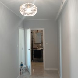 VPBud - Korytarz w odcieniach szarości z widokiem na łazienkę z czarną szafką pod umywalką i lustrem w złotej ramie, pod sufitem designerska lampa.